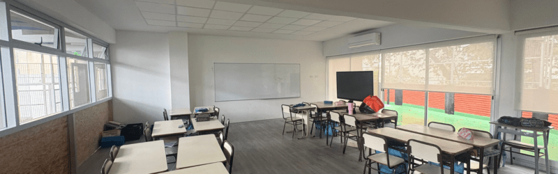 Aula renovada con ventanales amplios, cortinas roller, aire acondicionado y mobiliario escolar.